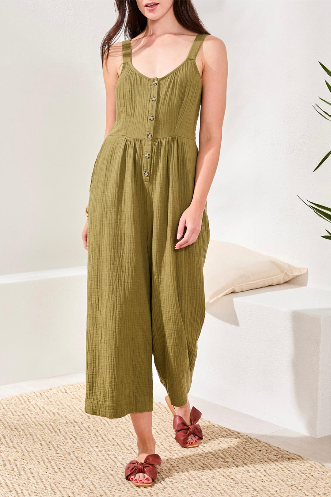 Woman wearing a green crinkle jumpsuit standing on a textured rug with a white wall background