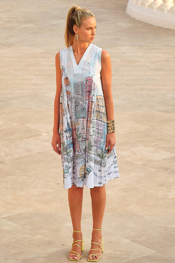 Woman wearing a sleeveless cotton v-neck dress with a cityscape print on a stone pavement.