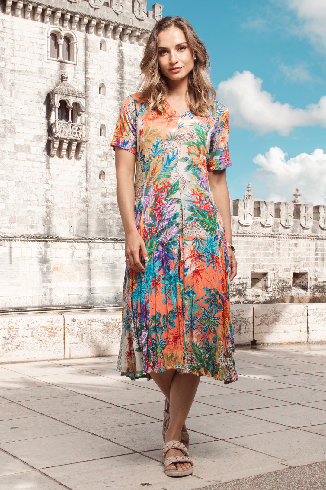 Woman in a colorful v-neck dress standing in front of a stone building with a blue sky.