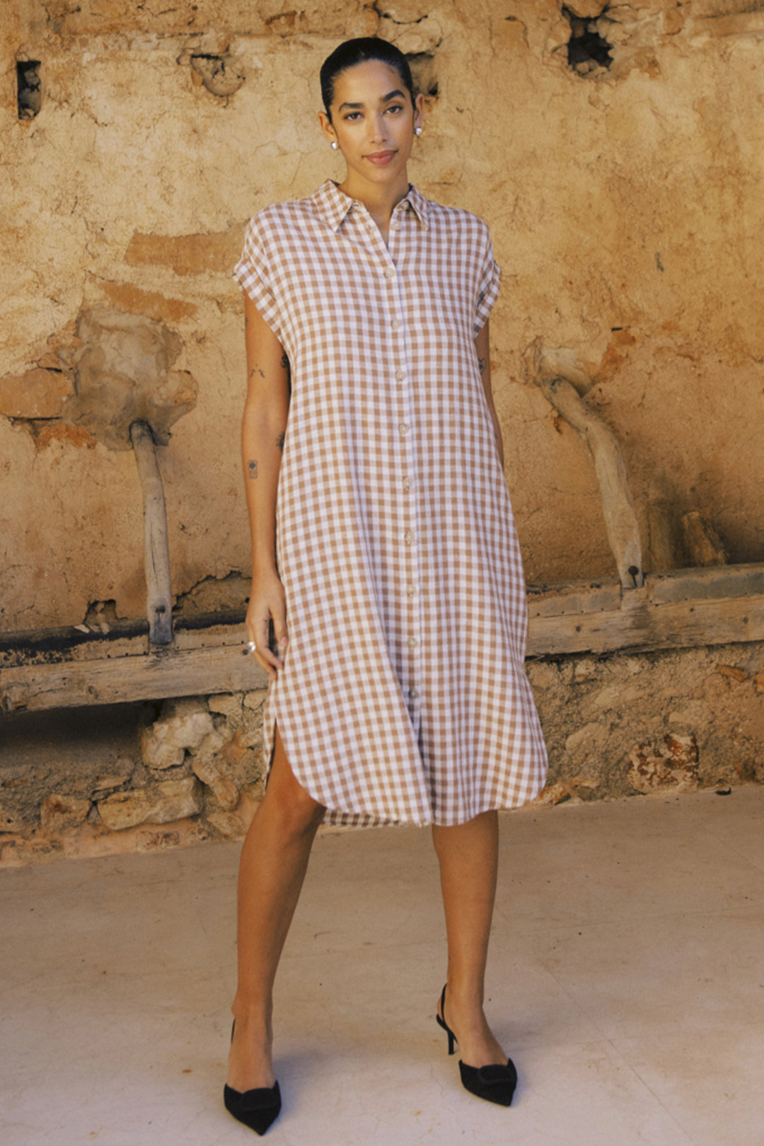 Woman wearing a checkered dress standing against a rustic wall.
