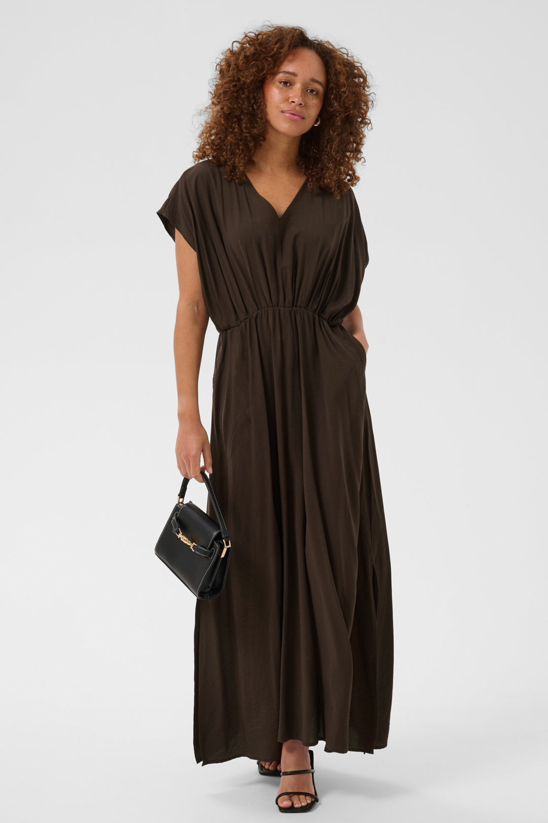 A woman modeling a dark brown, short-sleeved maxi dress with a V-neckline and gathered waist, holding a small black handbag against a light gray studio background.