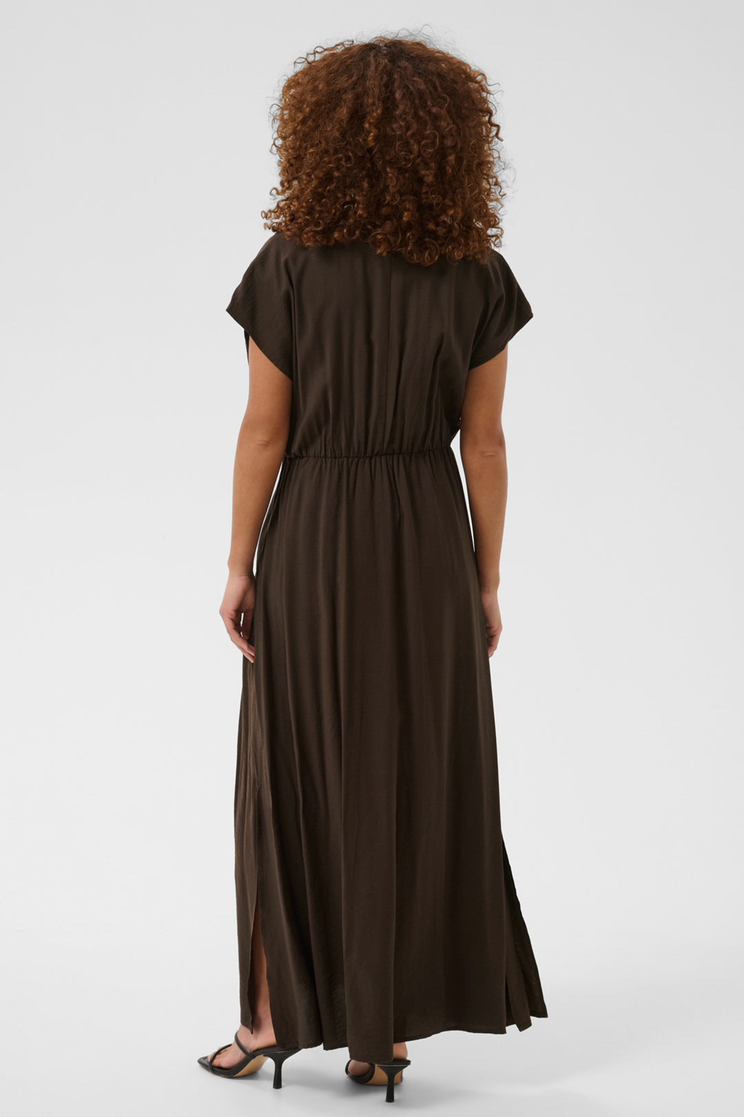 A woman with curly brown hair is shown from the back wearing a dark brown short-sleeve maxi dress with a gathered waist and side slits, paired with black heeled sandals, against a light gray studio background.
