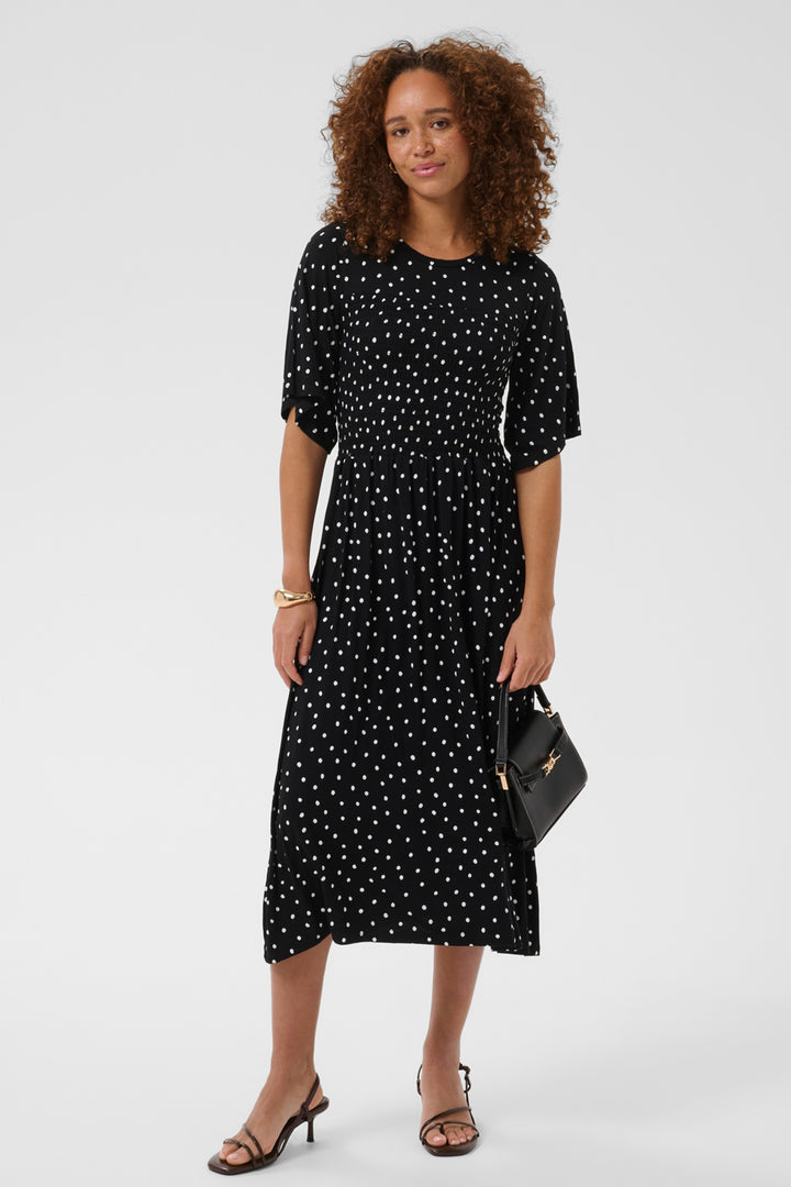 A woman with curly brown hair stands against a light gray studio background wearing a black midi dress with a white polka dot pattern, holding a small black handbag.