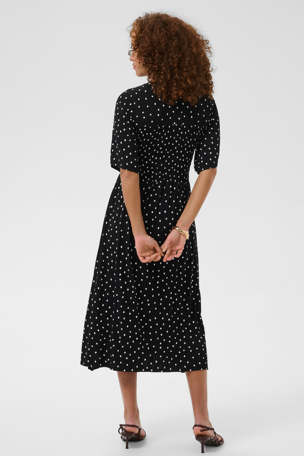 A woman with curly brown hair is shown from the back wearing a black midi dress with a white polka dot pattern, short puff sleeves, and a high collar. She is wearing black strappy heels against a plain light gray studio background.