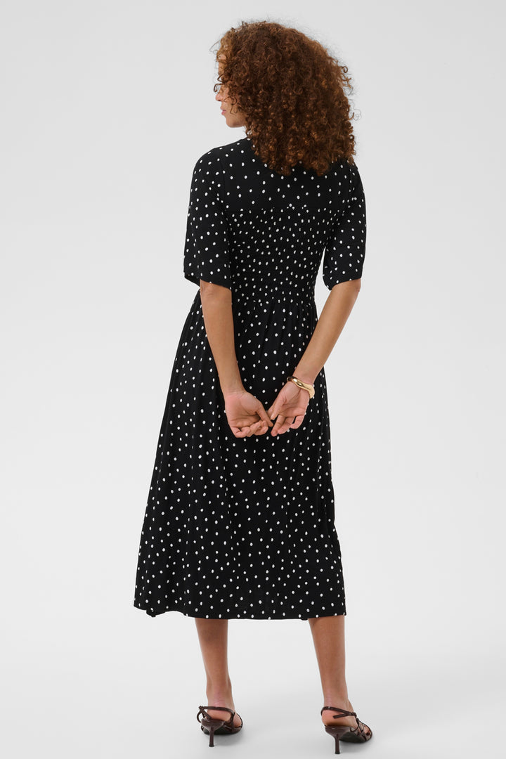 A woman with curly brown hair is shown from the back wearing a black midi dress with a white polka dot pattern, short puff sleeves, and a high collar. She is wearing black strappy heels against a plain light gray studio background.