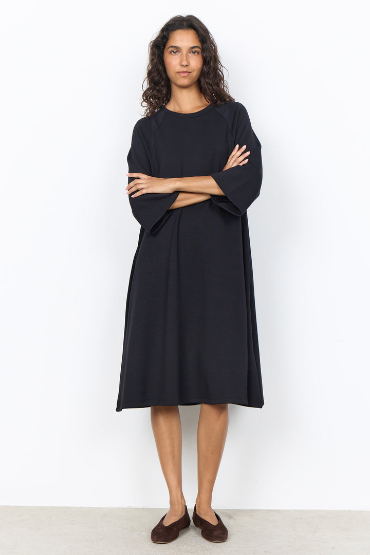 A woman modeling a loose black midi dress with wide three-quarter sleeves and a round neckline, paired with dark brown slip-on shoes, against a plain white studio background.