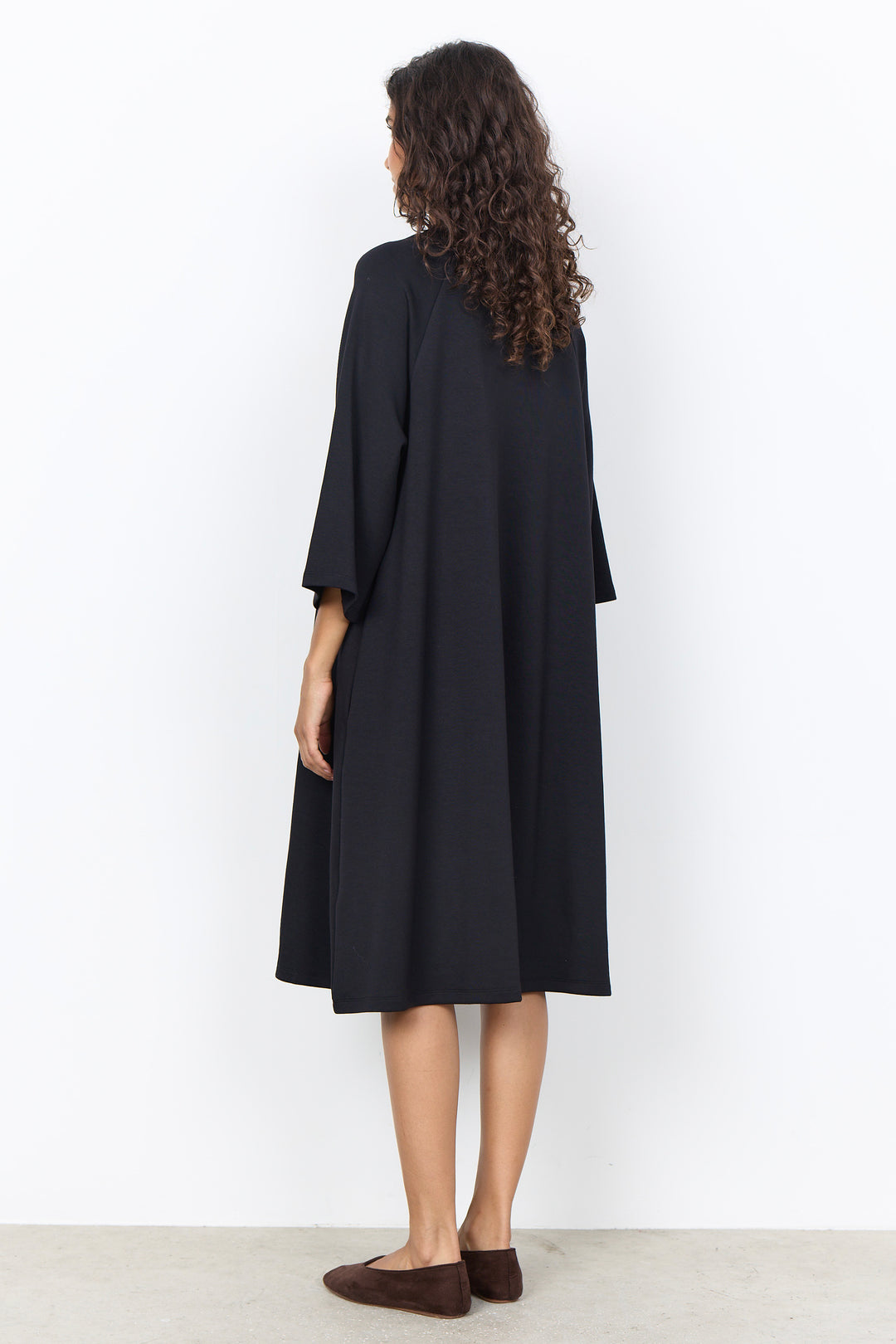 A woman with long curly brown hair is shown from the back wearing a loose, black midi-length tunic dress with wide three-quarter sleeves. She is standing against a plain white wall on a light gray floor.