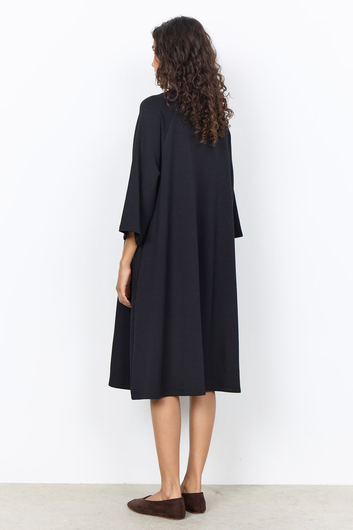 A woman with long curly brown hair is shown from the back wearing a loose, black midi-length tunic dress with wide three-quarter sleeves. She is standing against a plain white wall on a light gray floor.