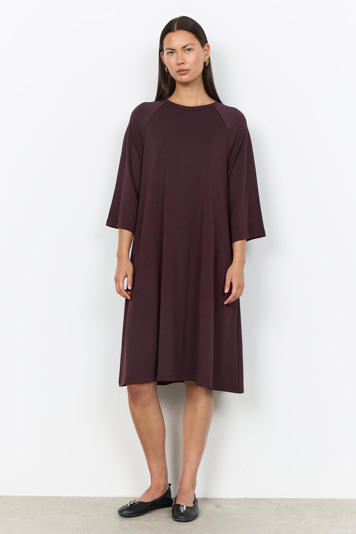 A woman modeling a dark plum-colored, knee-length dress with three-quarter raglan sleeves, standing against a plain white wall.