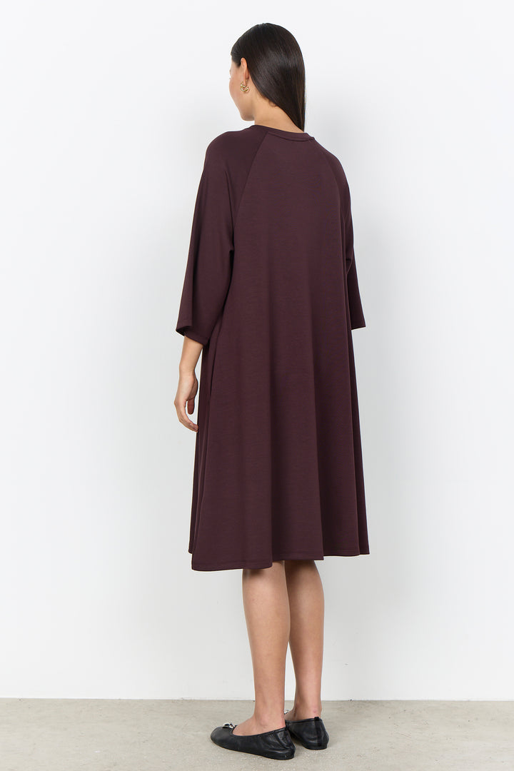 A woman viewed from the back wearing a loose, knee-length dark plum dress with three-quarter sleeves and black flats, standing against a plain white wall.