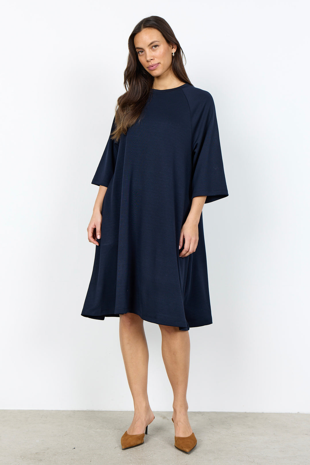 A woman modeling a navy blue shift dress with three-quarter sleeves and a relaxed silhouette, paired with brown pointed-toe heels, against a plain white studio background.