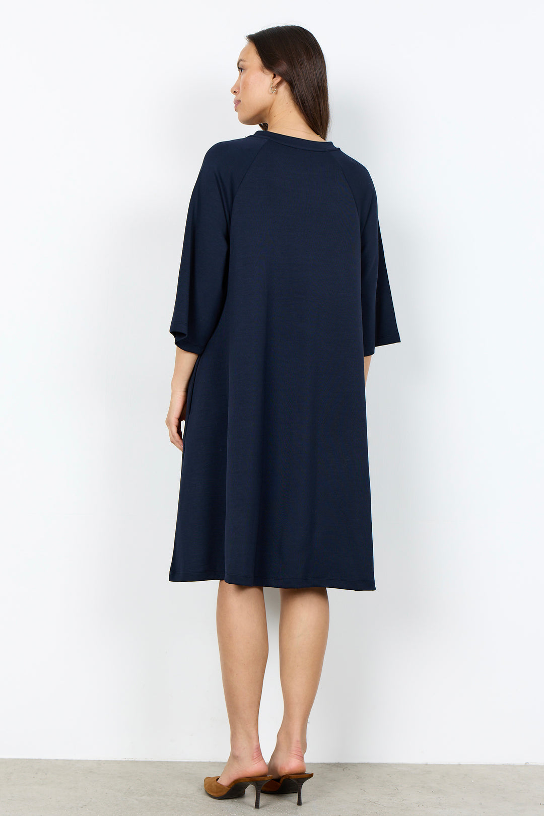 A woman modeling a navy blue shift dress with three-quarter sleeves and a relaxed silhouette, paired with brown pointed-toe heels, against a plain white studio background.