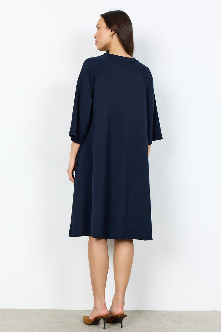A woman modeling a navy blue shift dress with three-quarter sleeves and a relaxed silhouette, paired with brown pointed-toe heels, against a plain white studio background.