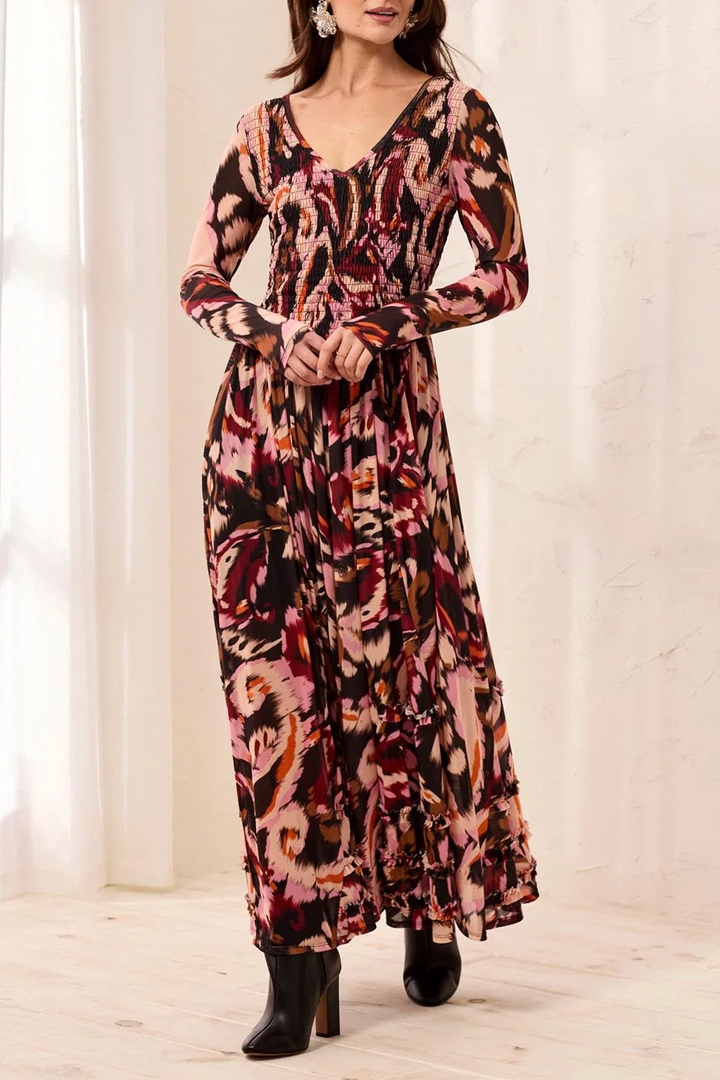 A woman modeling a long-sleeve maxi dress with a multicolor abstract floral print in pink, red, orange, and brown tones, photographed in a bright indoor setting with white curtains.