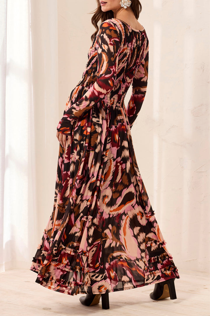 A woman modeling a long-sleeve maxi dress with a multicolor abstract floral print in pink, red, orange, and brown tones, photographed in a bright indoor setting with white curtains.