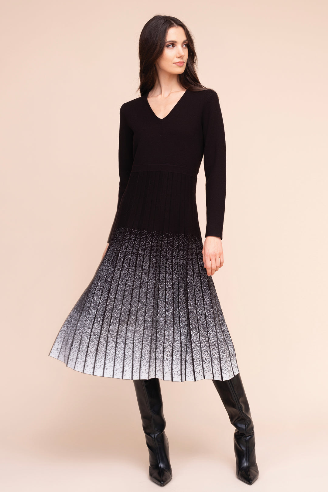 A woman modeling a long-sleeve black knit dress with a pleated skirt that fades from black at the top to white at the hem, paired with black knee-high boots, against a solid beige studio background.