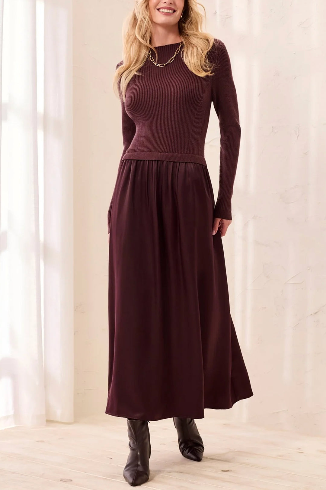 A woman modelling a brown long sleeve maxi dress standing in front of a beige curtain background. 