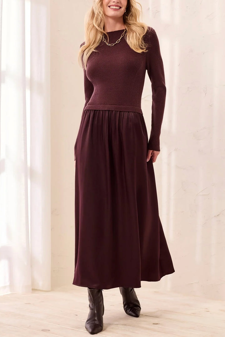 A woman modelling a brown long sleeve maxi dress standing in front of a beige curtain background. 