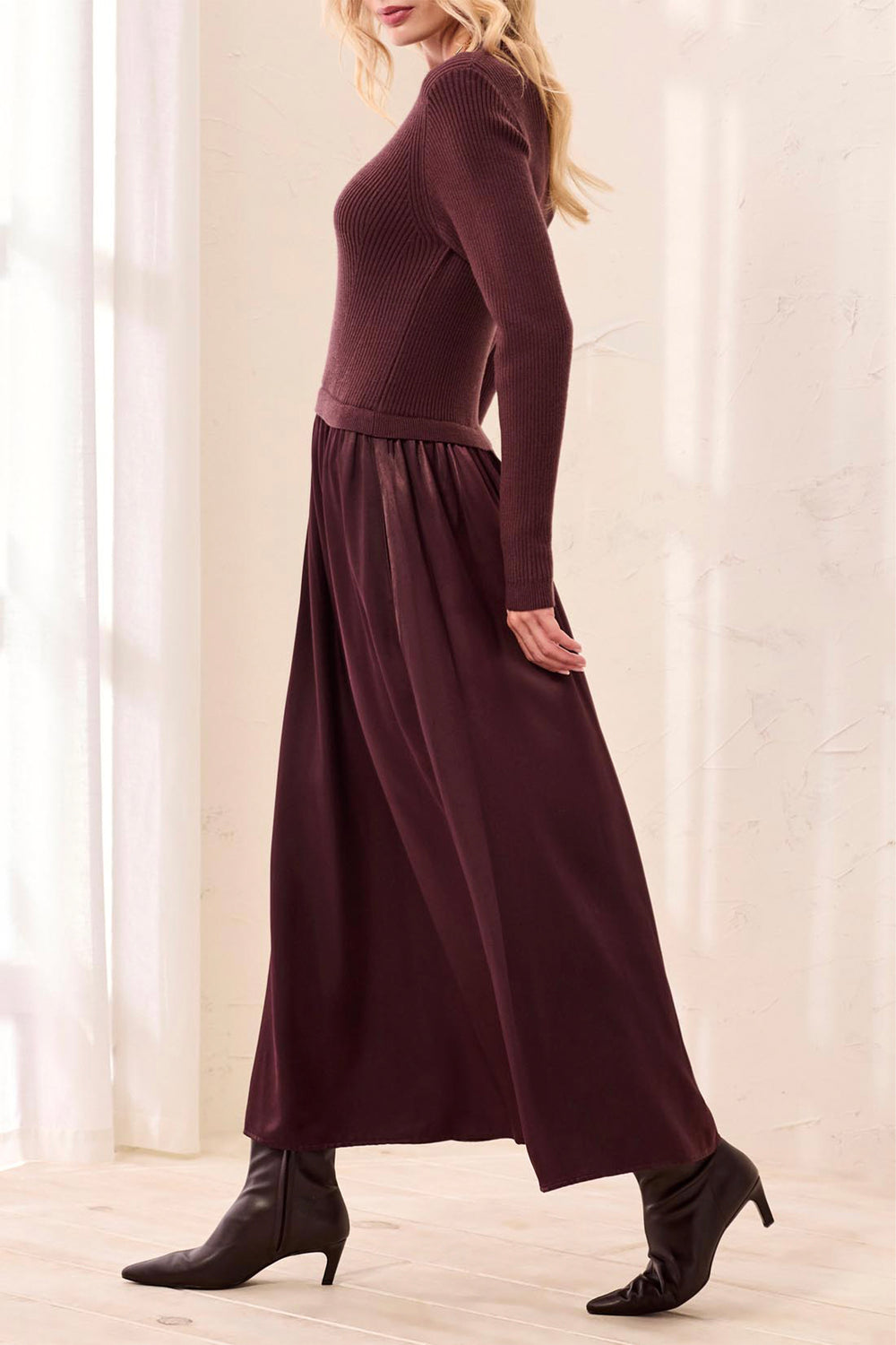 A woman shown from the side modelling a brown long sleeve maxi dress standing in front of a beige curtain background. 
