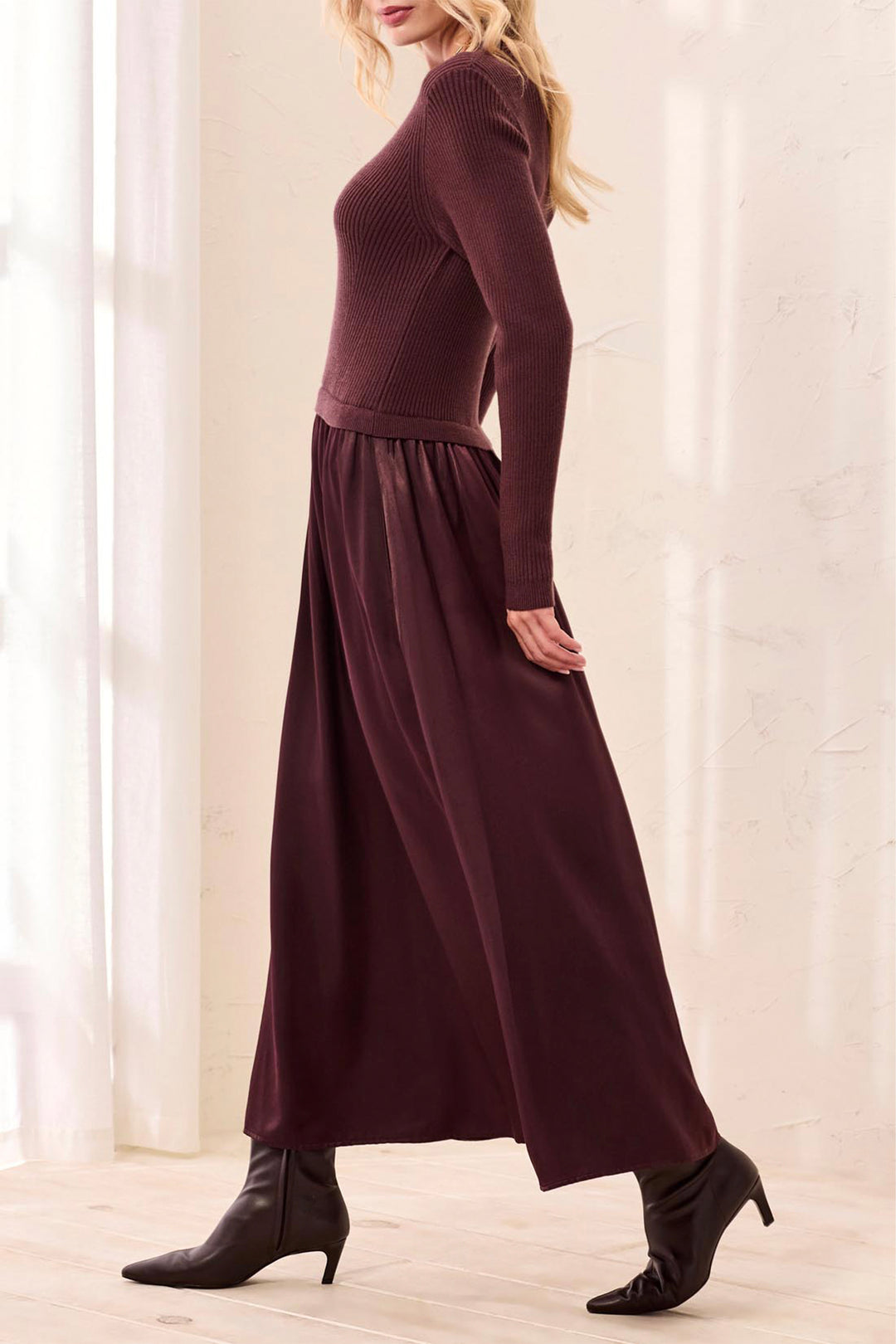 A woman shown from the side modelling a brown long sleeve maxi dress standing in front of a beige curtain background. 