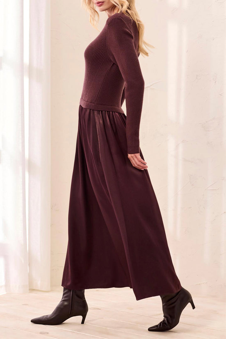 A woman shown from the side modelling a brown long sleeve maxi dress standing in front of a beige curtain background. 