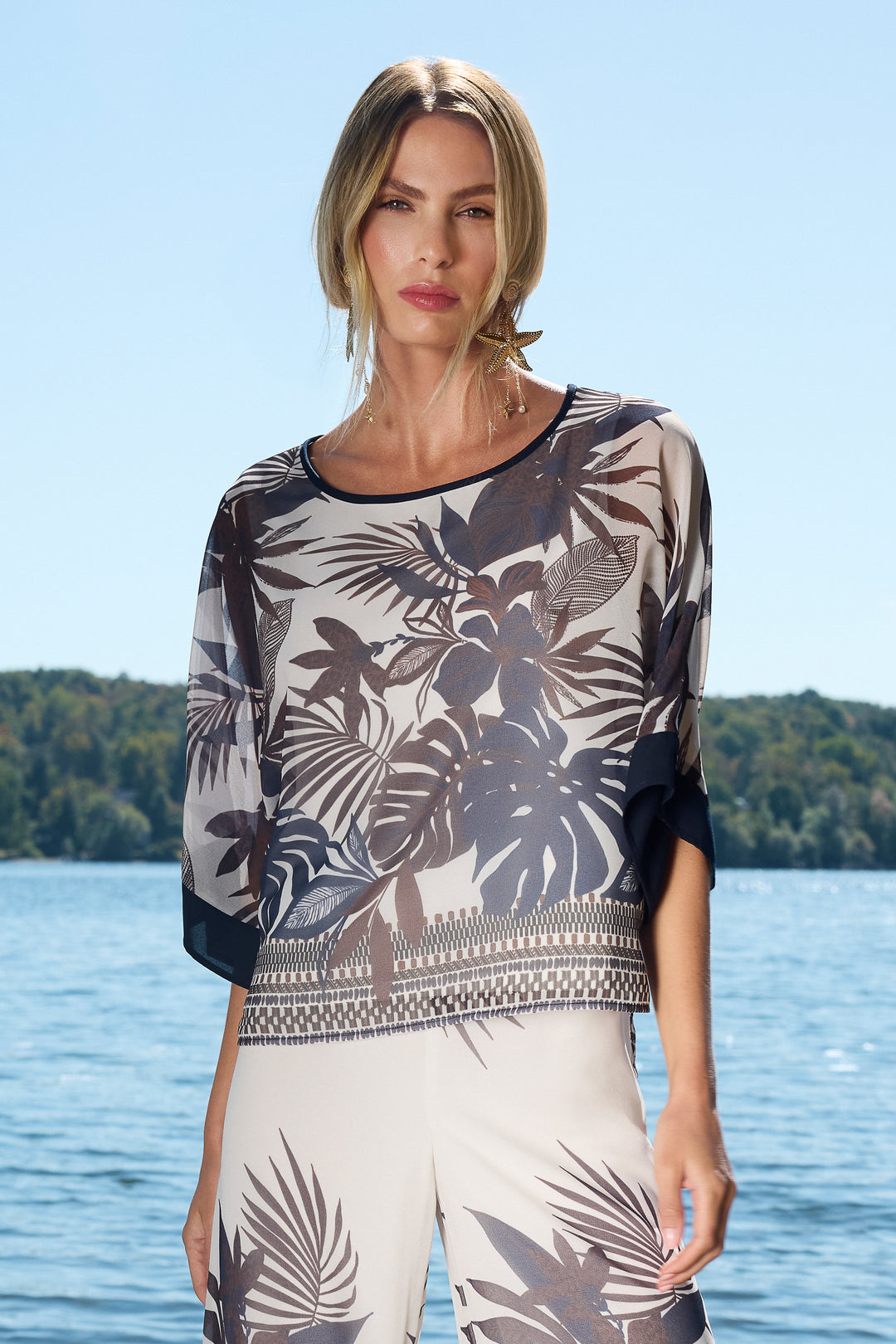 Woman wearing a patterned floral top and white pants by a lake