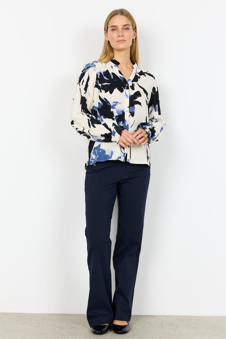 Woman wearing a patterned blouse and navy pants against a white background