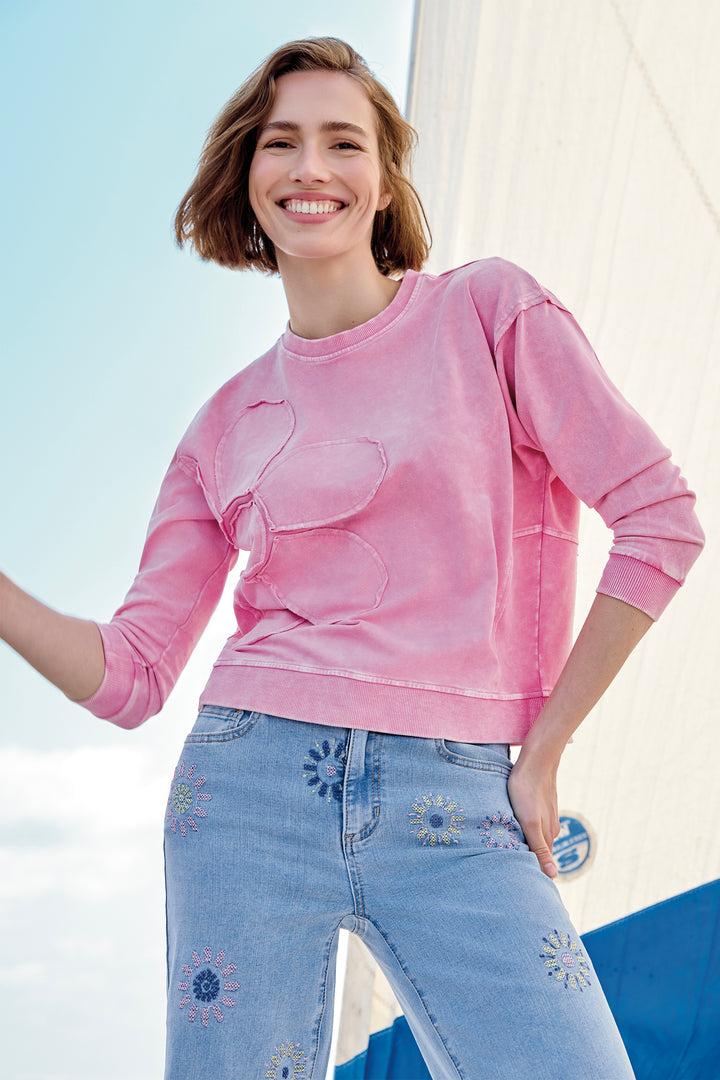 Woman wearing a pink sweatshirt with a heart design and blue jeans, standing against a light blue sky.