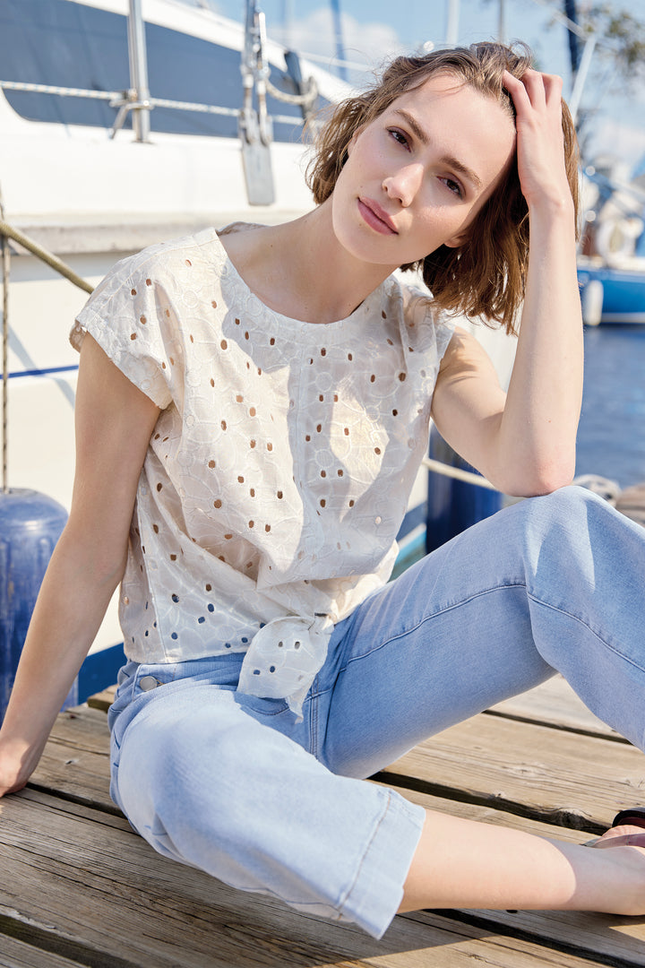 Woman sitting on a dock wearing a white blouse and light blue jeans, with boats in the background.