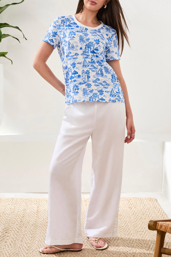 Woman wearing a blue and white patterned top and white pants in a bright room.