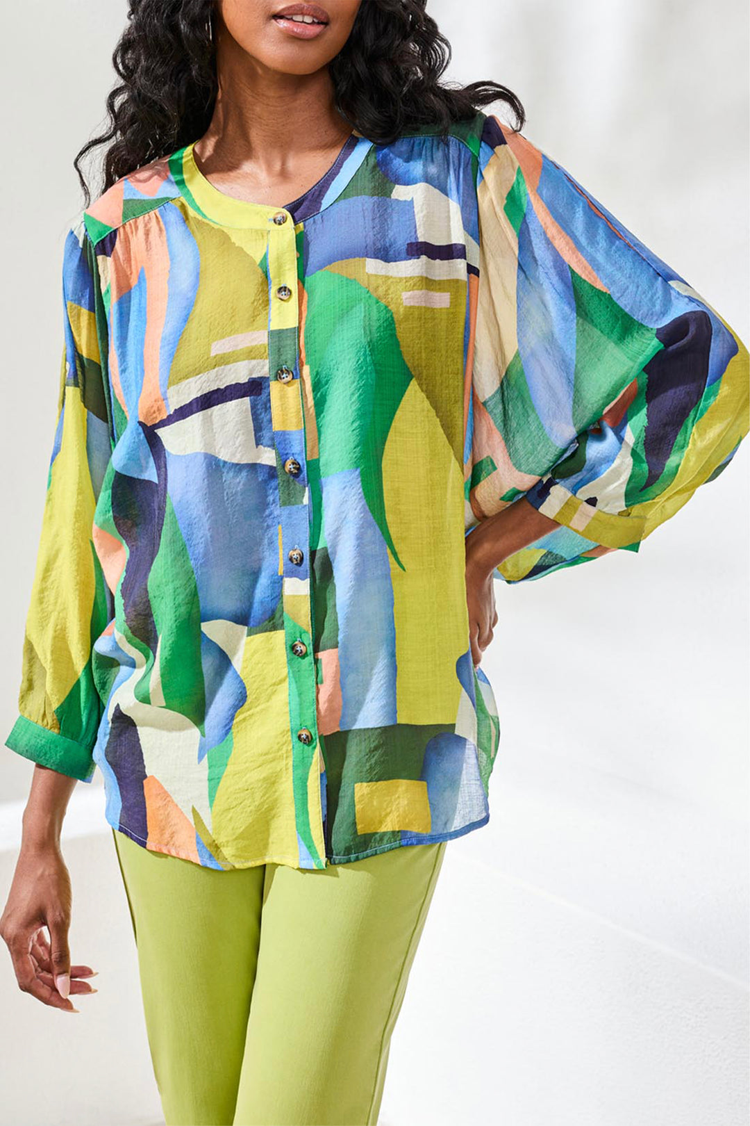 Woman wearing a colorful patterned button top and green pants on a white background