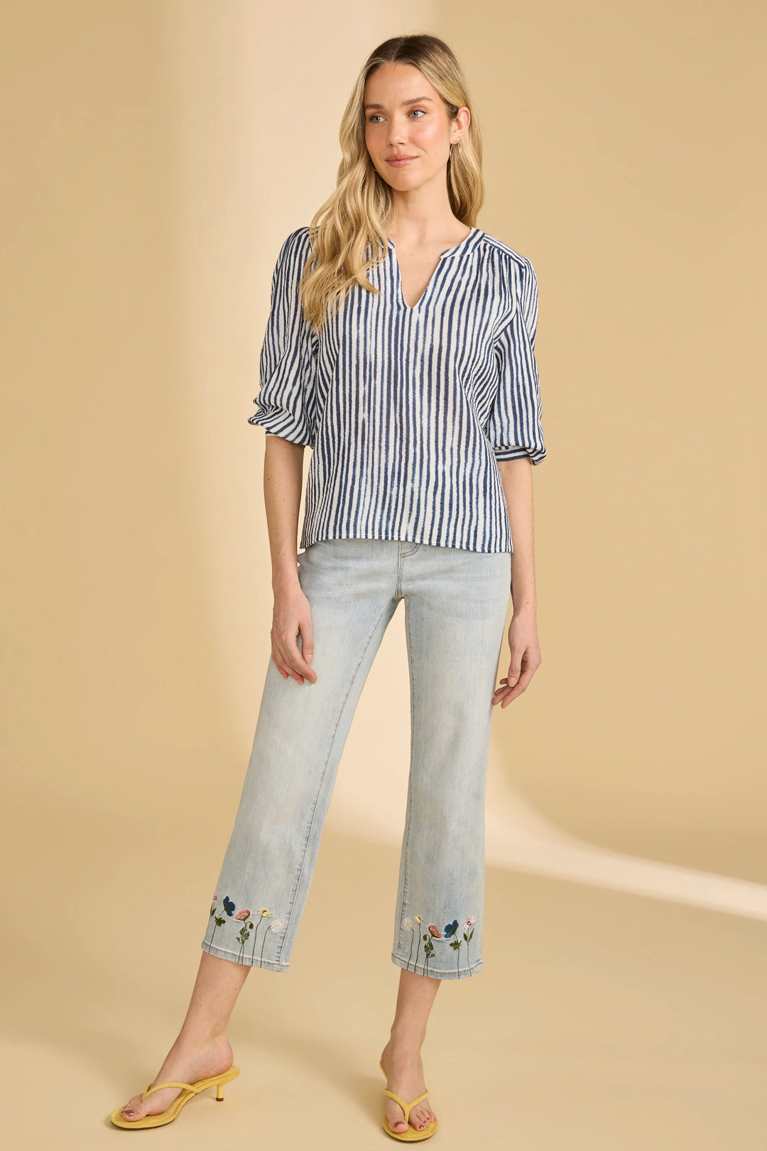 Woman wearing a blue and white striped shirt and light blue jeans with floral embroidery on a beige background