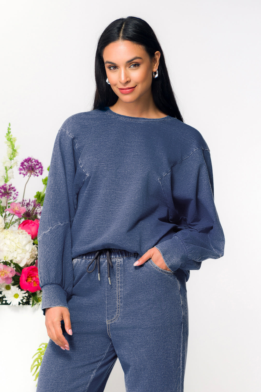 Woman wearing a blue sweater and jeans with a white background and floral arrangement.