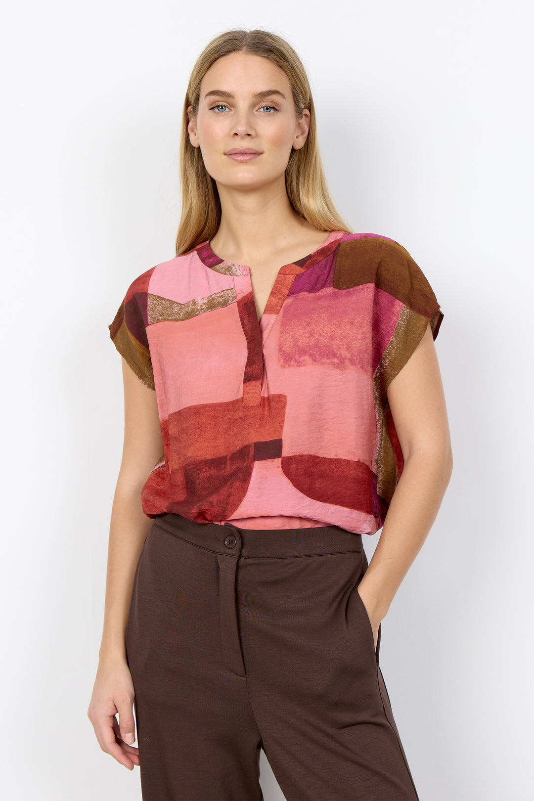 Woman wearing a colorful patterned top  with split neck and brown pants on a white background