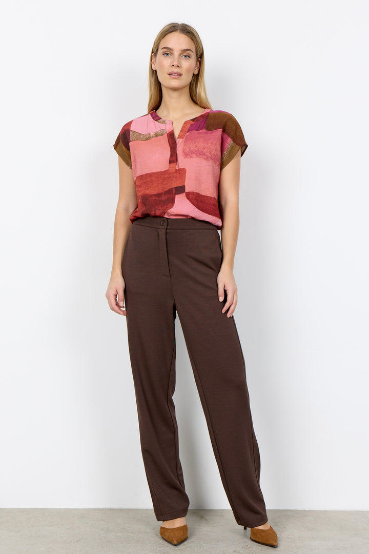 Woman wearing a patterned top and brown pants against a white background