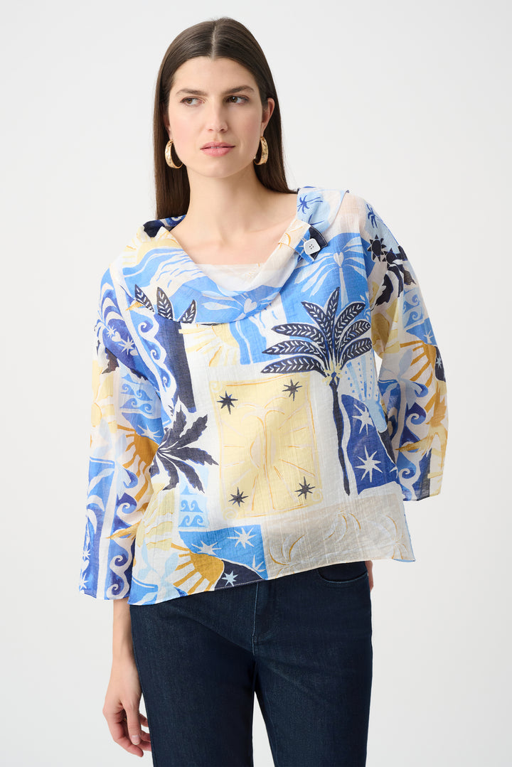 A woman modeling a lightweight, semi-sheer three-quarter sleeve top with a colorful patchwork print of blue, yellow, and white tropical and abstract motifs, paired with dark blue jeans against a plain light gray background.