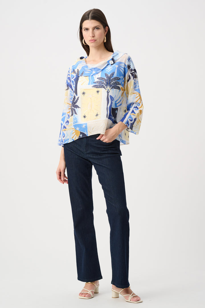 A woman modeling a tropical print blouse with blue, yellow, and white palm tree patterns, paired with dark blue jeans and beige sandals, against a light gray studio background.