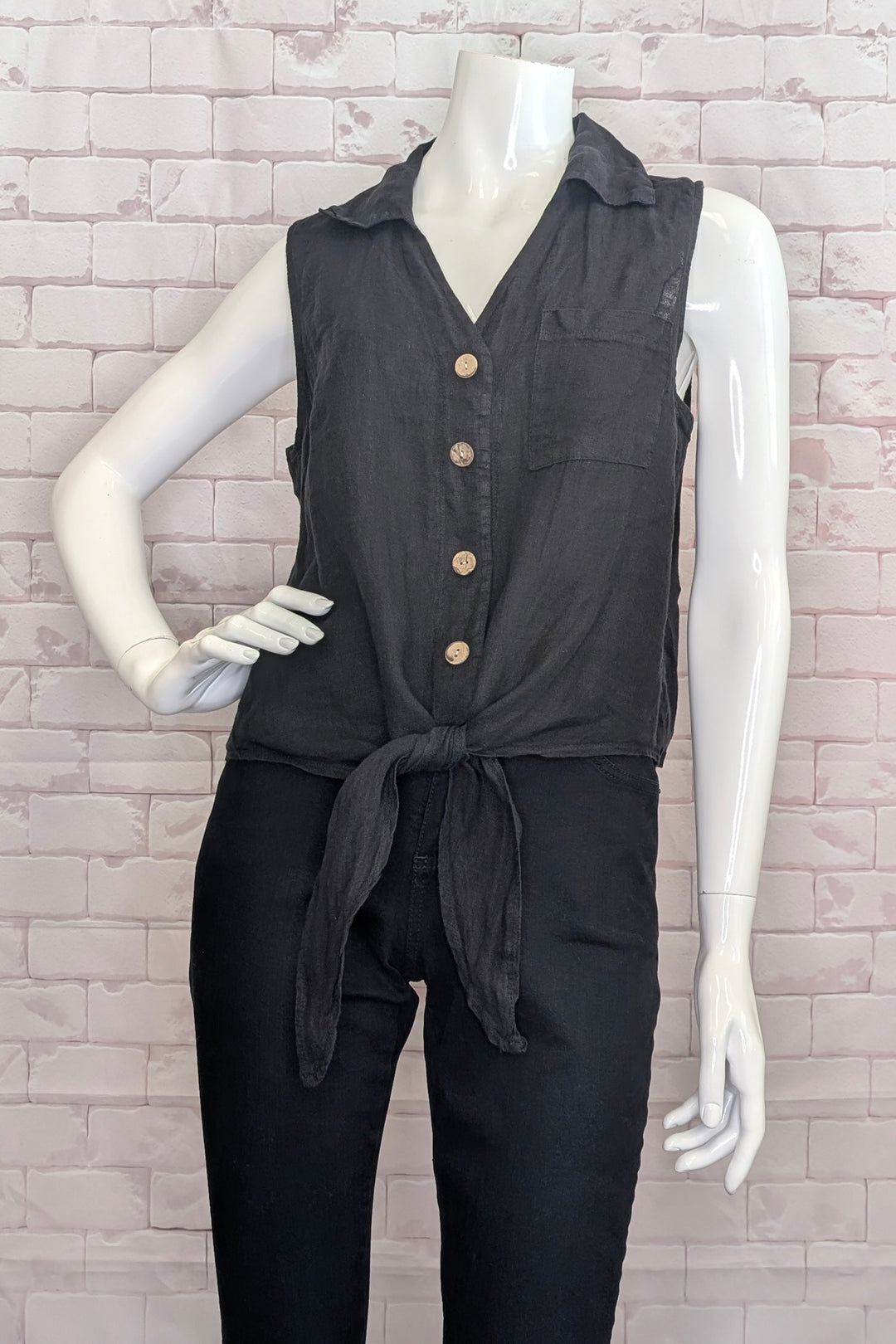 Mannequin wearing a black sleeveless top with buttons and matching pants against a light brick wall.