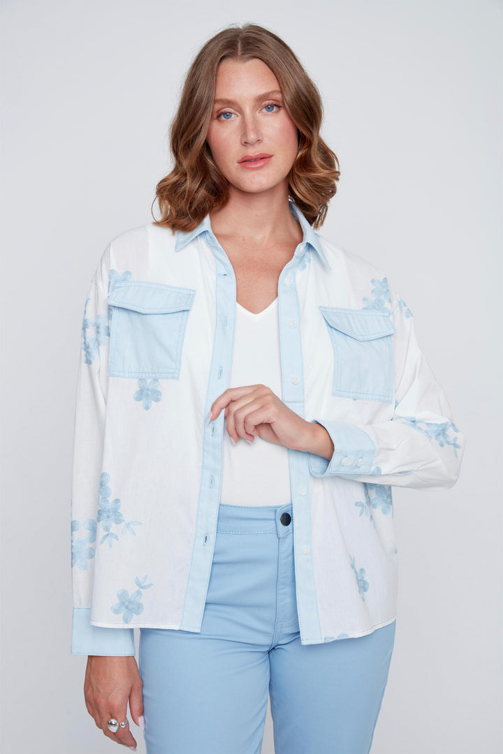 Woman wearing a light blue and white floral shirt over a white top with light blue jeans on a plain background