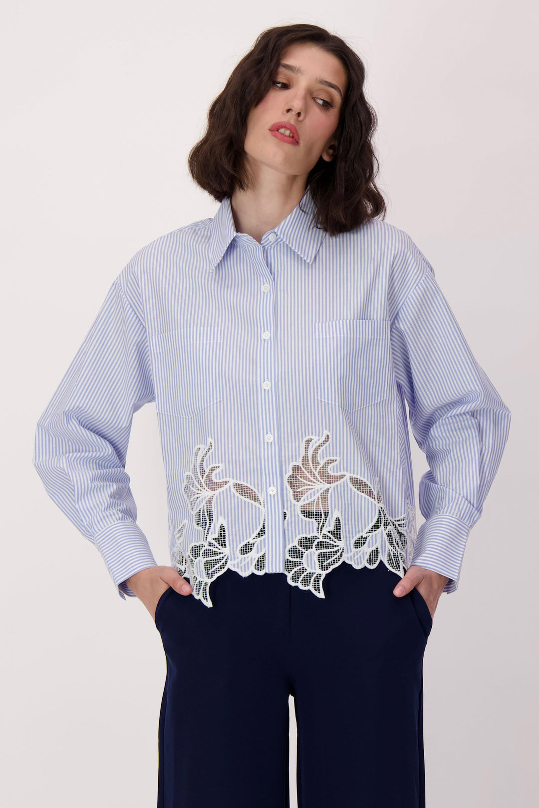 Woman wearing a light blue stripe shirt with floral lace details along hem on a white background