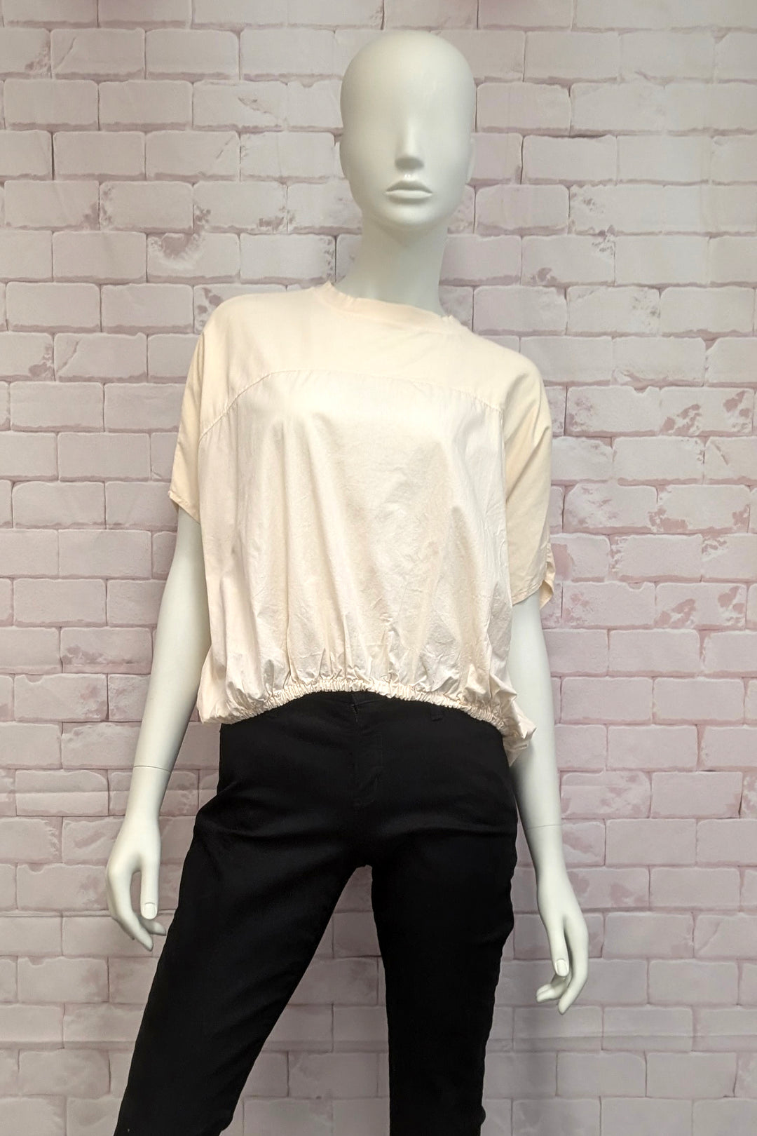 Mannequin wearing a cotton beige blouse and black pants against a light brick wall.
