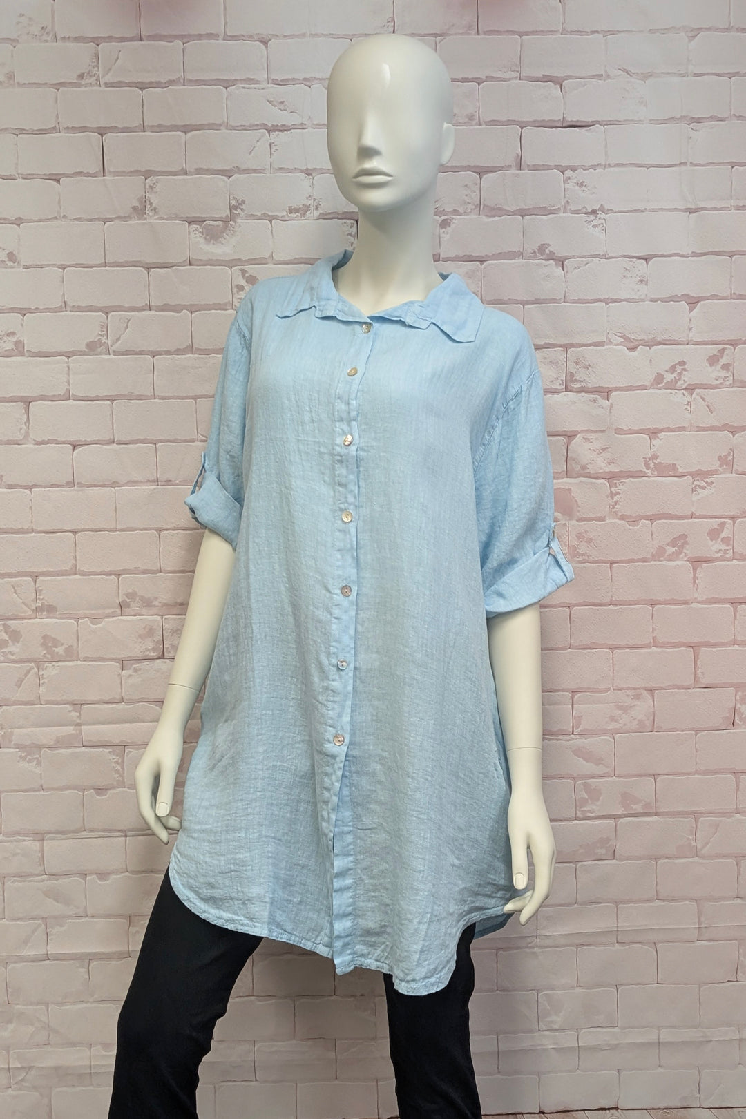 Mannequin wearing a light blue button-up tunic linen shirt against a brick wall.