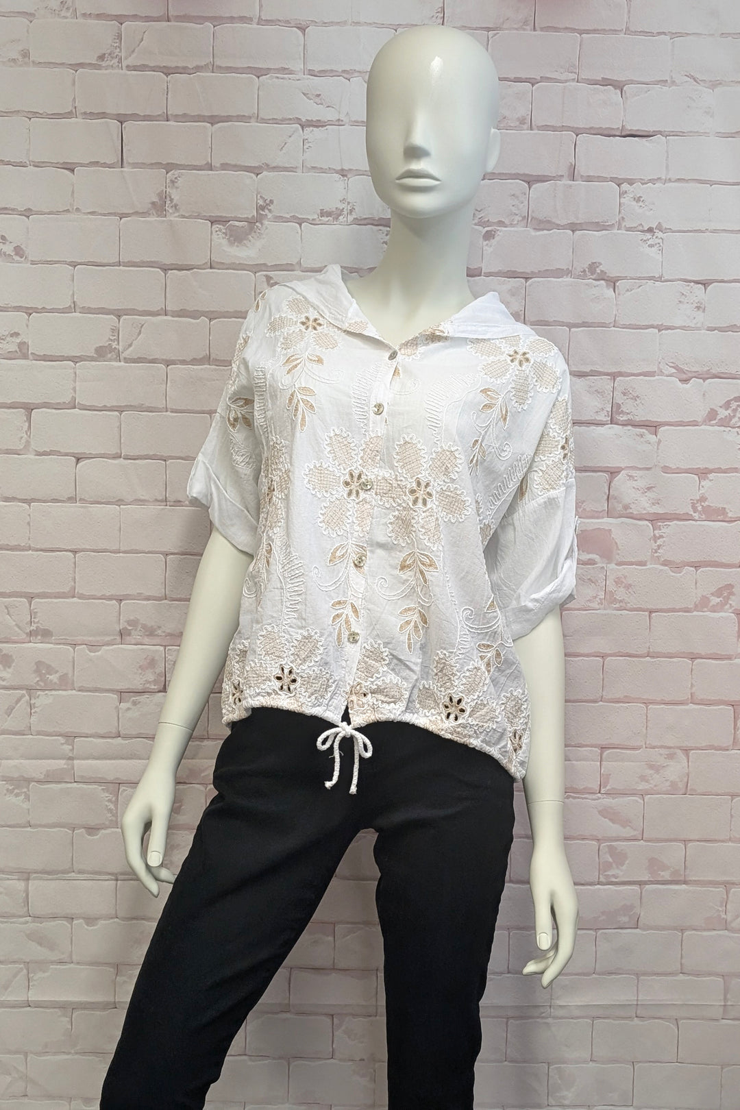 Mannequin wearing a white lace floral top and black pants against a light brick wall.