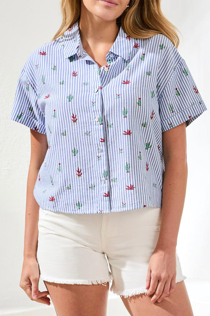 Woman wearing a blue and white striped shirt with cactus pattern, short sleeves and white shorts on a plain background