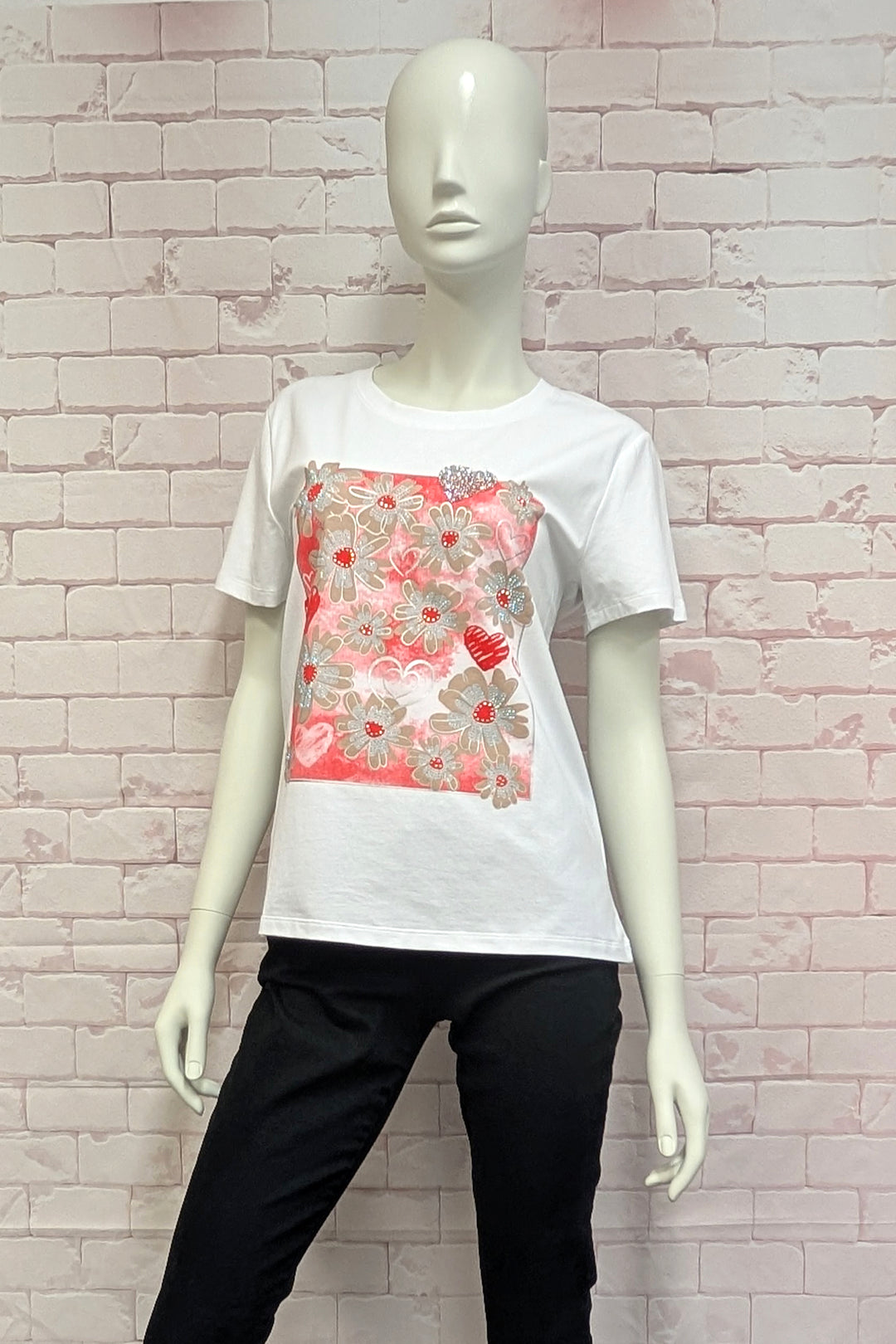 Mannequin wearing a white t-shirt with a floral design and black pants against a light brick wall.