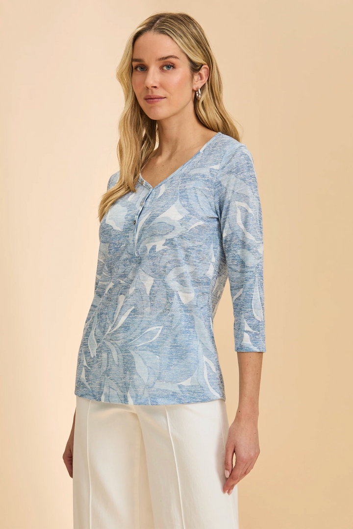 Woman wearing a blue patterned notch v-neck top with 3/4 sleeves and white pants on a beige background