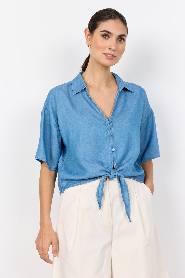 Woman wearing a blue button-up shirt tied at the waist with white pants on a white background