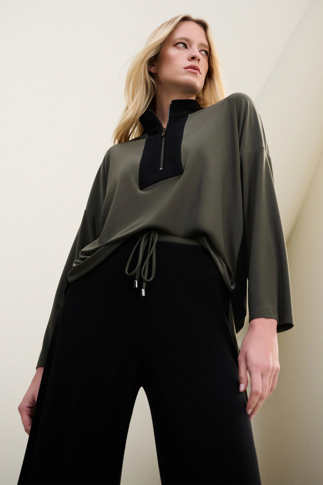Woman wearing a green zipper top with black accents and black pants against a plain background