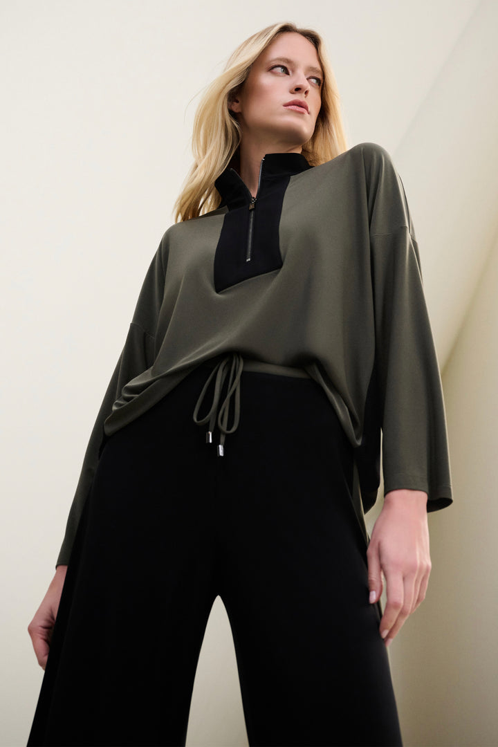 Woman wearing a green zipper top with black accents and black pants against a plain background