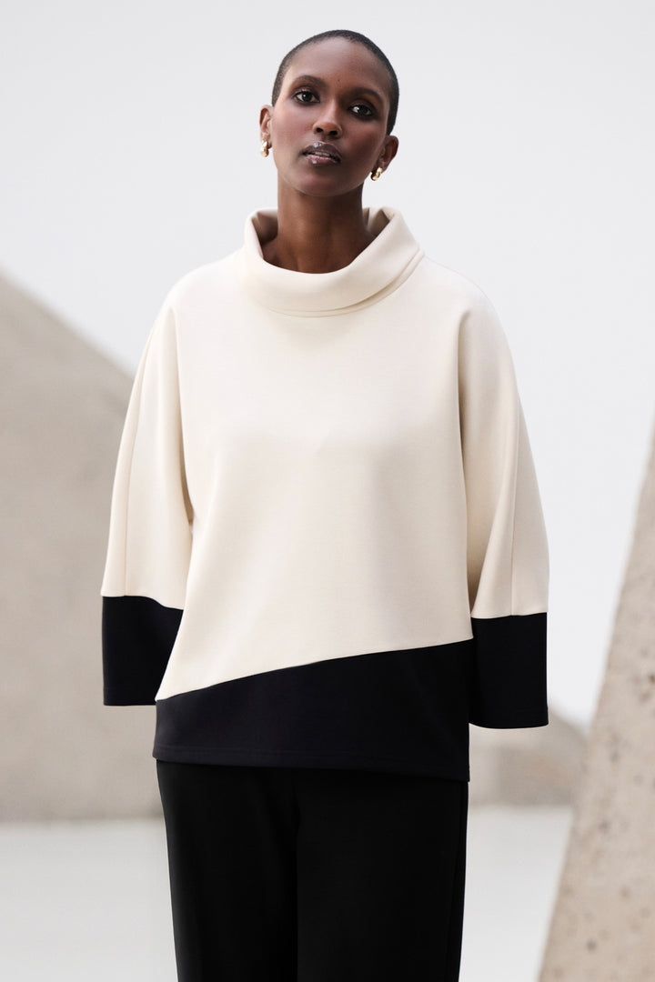 Woman wearing a cream and black pullover cowl neck top with a plain background
